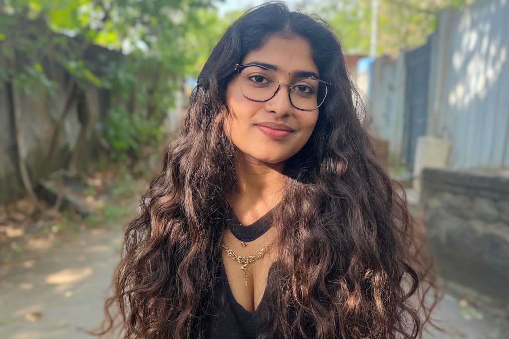 Source image of a young South Asian woman with long wavy hair and glasses outdoors, used by Dzinepixel to compare AI image-upscaling results from Nano Banana 2 Lite, Grok Imagine, ChatGPT 1.5, and FLUX 2 Pro